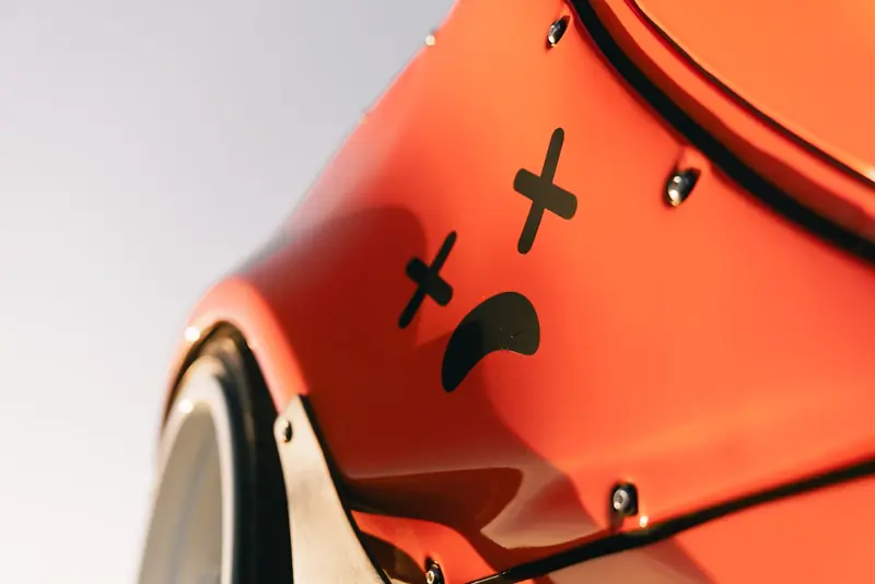 Close-up of an orange car's body featuring a stylized face decal under warm sunlight