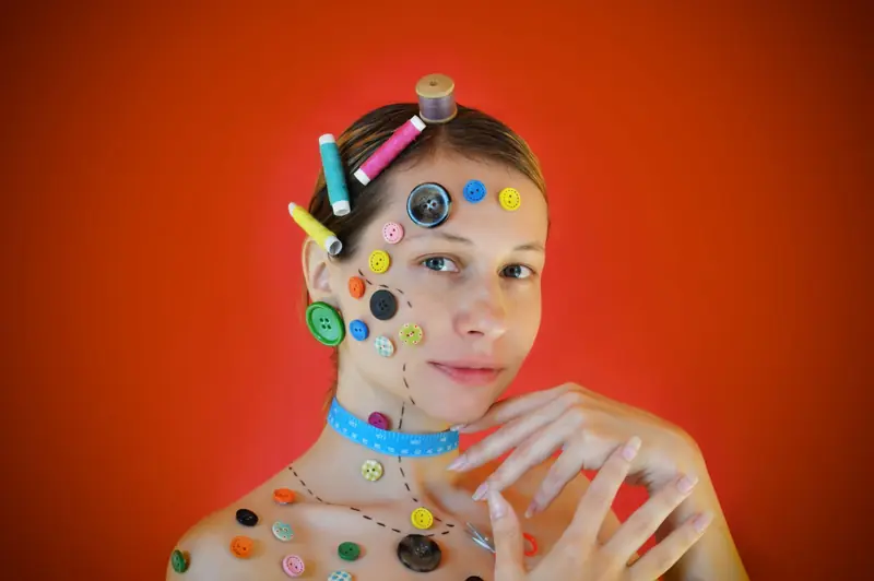 Young attractive female model with tailors meters around neck and colorful buttons and sewing spools on skin and hair touching face and looking at camera in stu