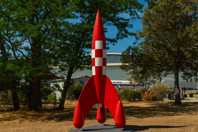 A striking red model rocket displayed outdoors in a sunny park setting in Toulouse, France