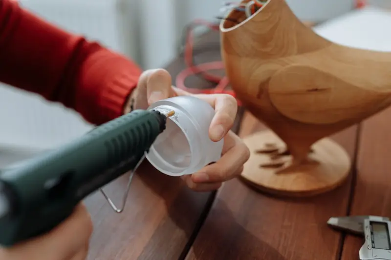 Close-up of a person gluing a plastic lid next to a wooden chicken model on a table