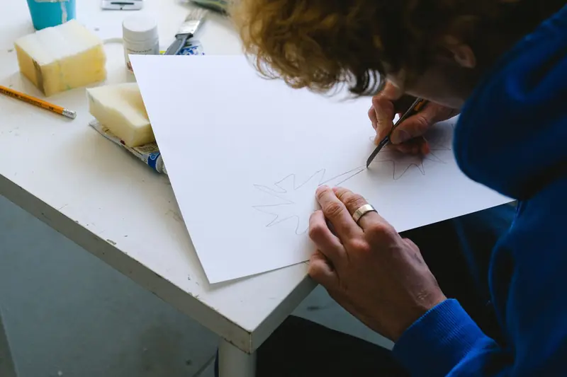 High angle of crop unrecognizable man in casual clothes sitting at table while cutting sketch on paper with utility knife near paints and sponge with pencil in 